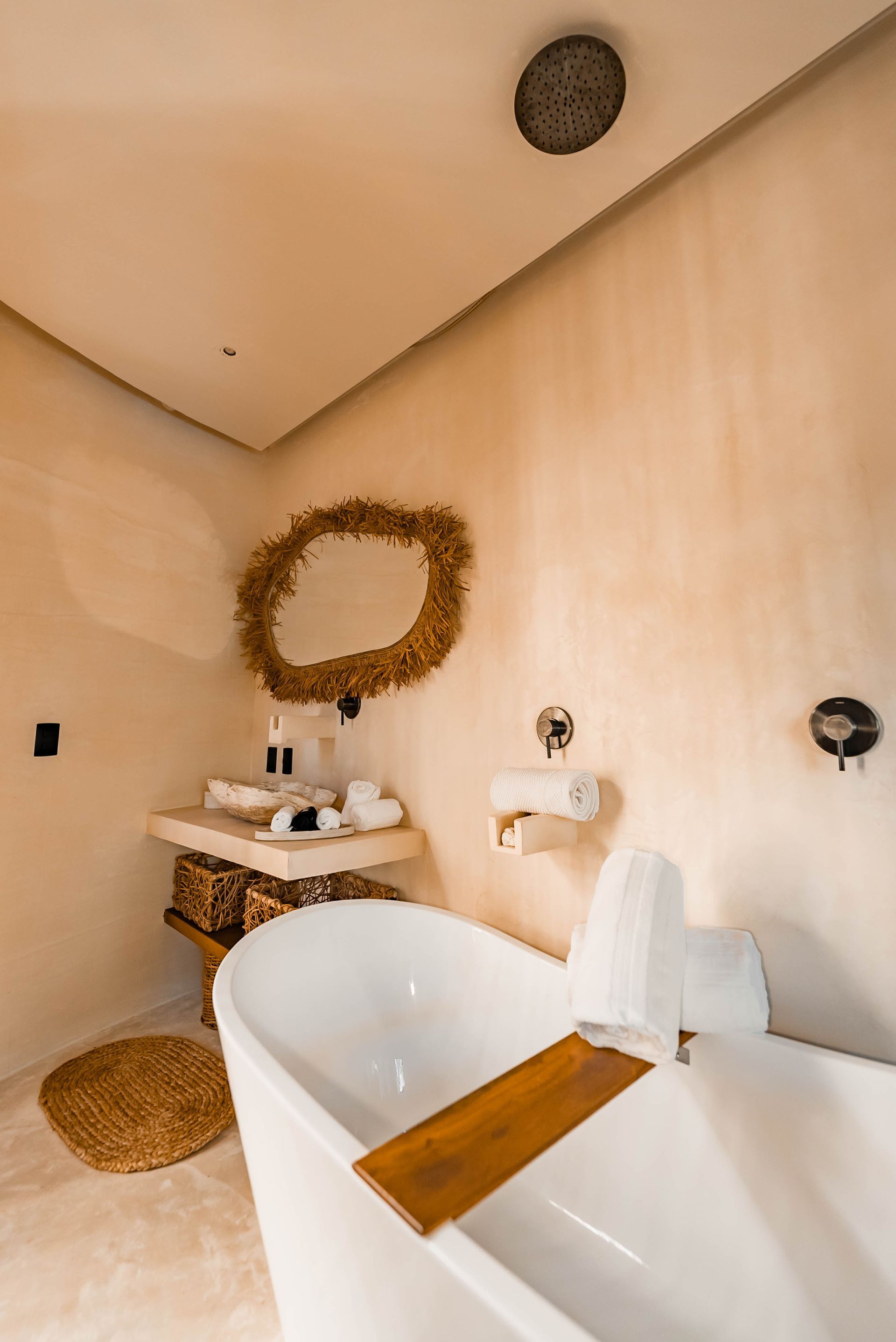 A minimalist bathroom with a white freestanding tub, a wooden caddy, a round textured mirror, and neutral walls.