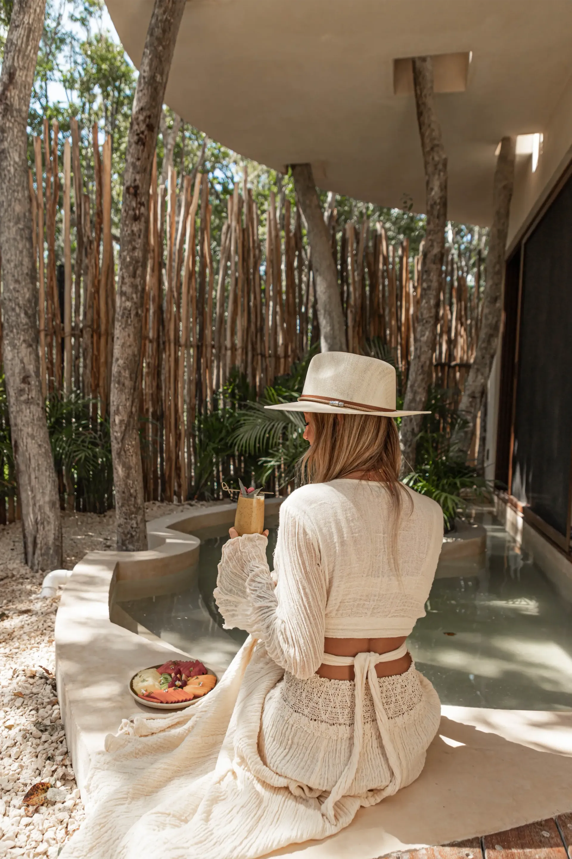 A person in a crochet outfit and wide-brim hat sits by a small pool in a tropical, wooded setting with a drink.