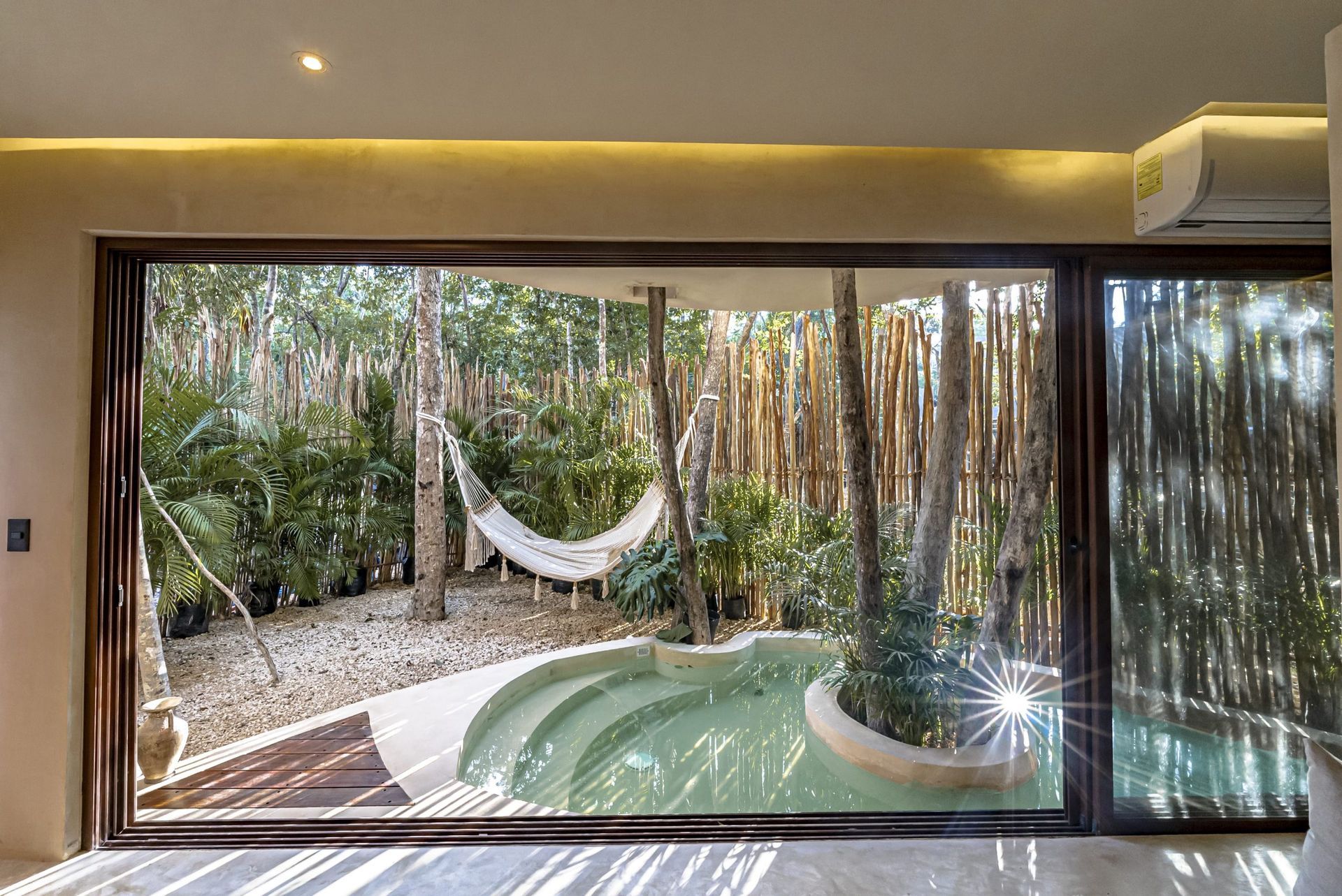 A view through a large glass door into a private courtyard featuring a small wading pool, a hammock, and lush trees.