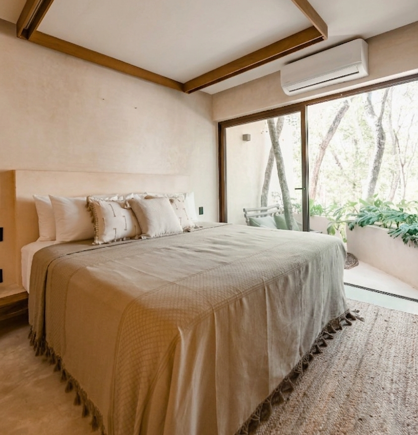 Bright bedroom with a bed, neutral decor, wood beams, and a sliding glass door to a balcony.