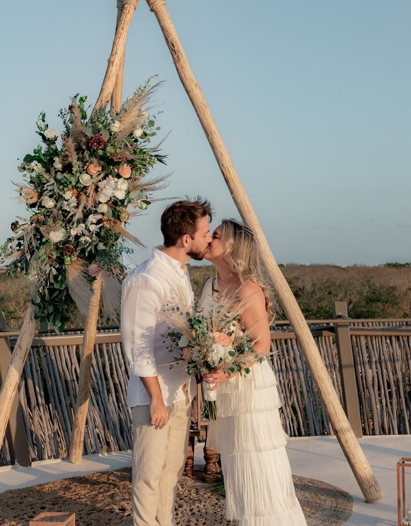 Couple kissing under a floral wedding arch at sunset on a rooftop overlooking water
