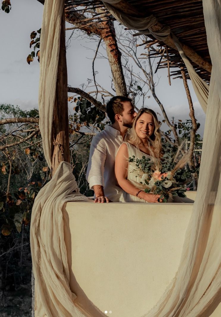 Couple posing on a balcony, holding a bouquet, framed by flowing drapes at sunset.
