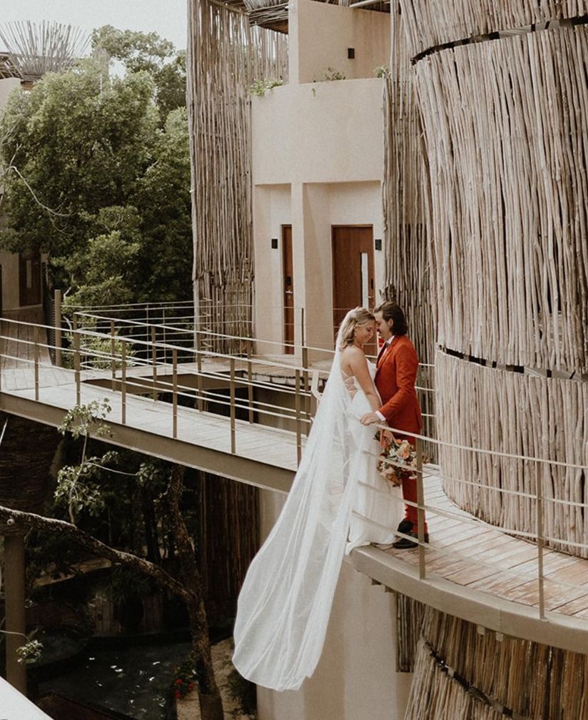 Bride and groom standing on a curved balcony walkway, embracing near a waterfall and trees