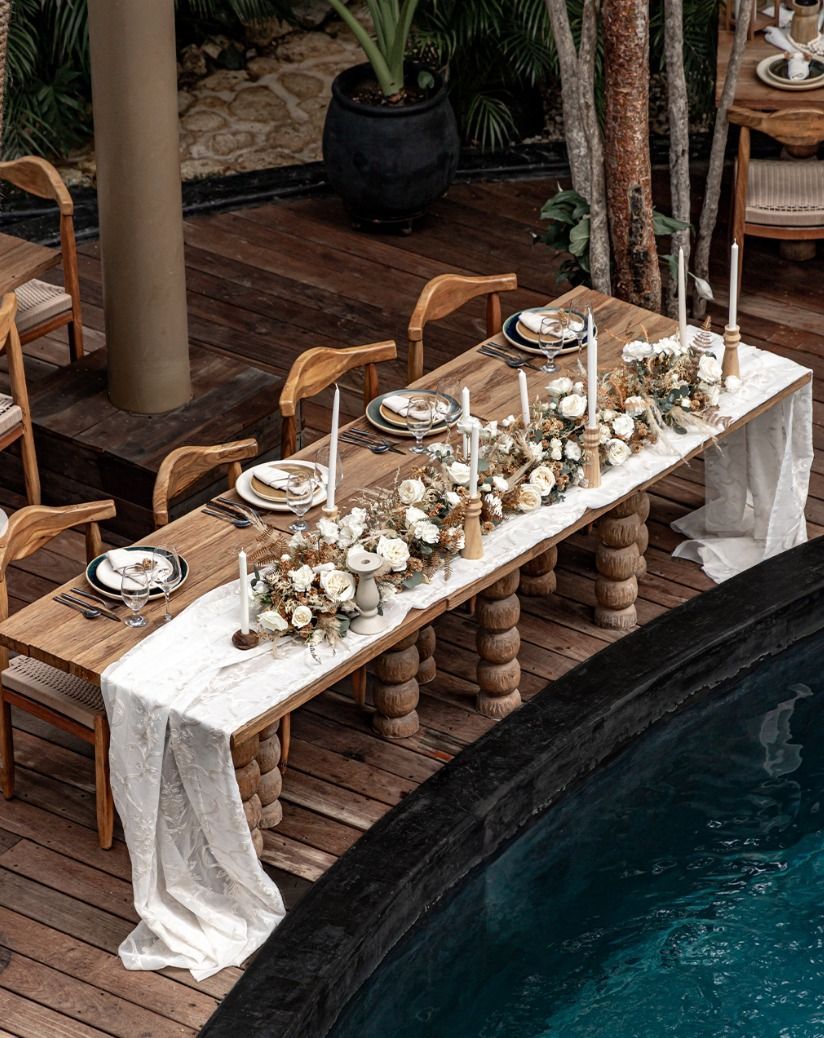 Long outdoor dining table by a pool, set with white linens, candles, and small floral arrangements