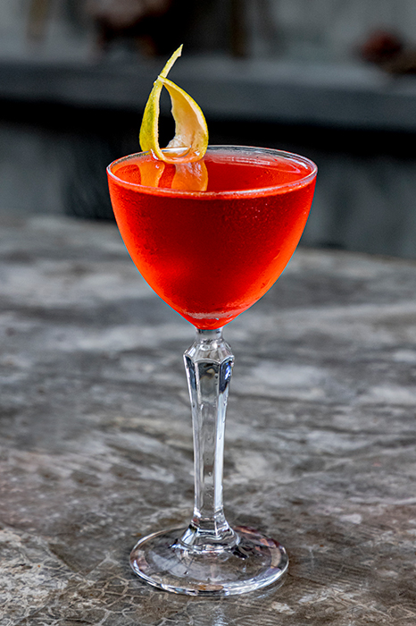A bright red cocktail in a stemmed coupe glass, garnished with a citrus peel twist, sitting on a stone surface.