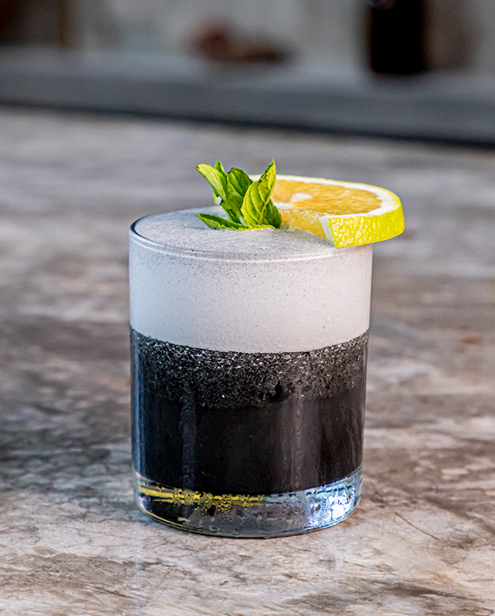 A dark, charcoal-colored cocktail topped with a thick white foam, garnished with a lemon slice and mint on a stone surface.