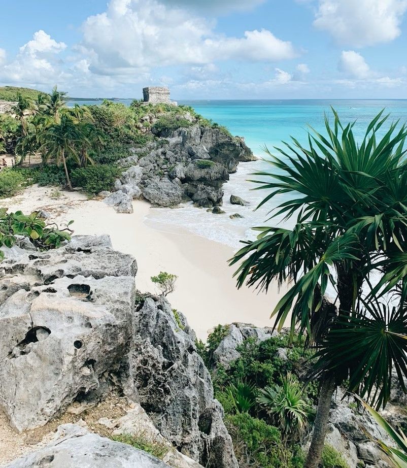 Tropical beach with white sand, rocky cliffs, turquoise water, and palm trees under a partly cloudy sky