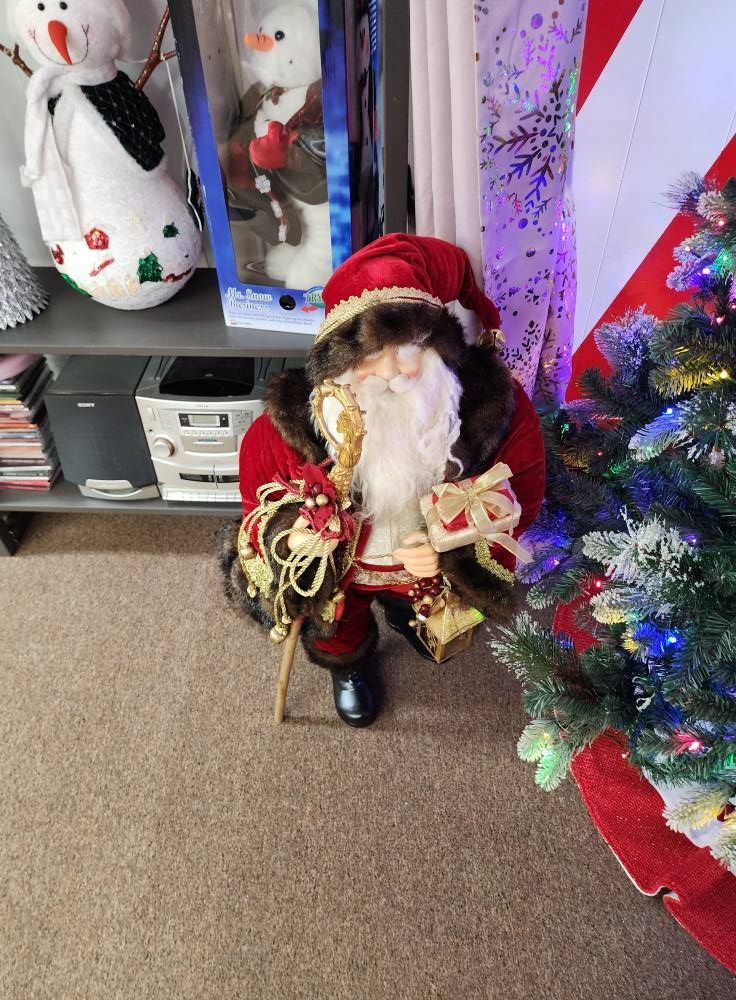 Santa Claus figure holding presents, next to Christmas tree. Red and gold decorations in a festive setting.