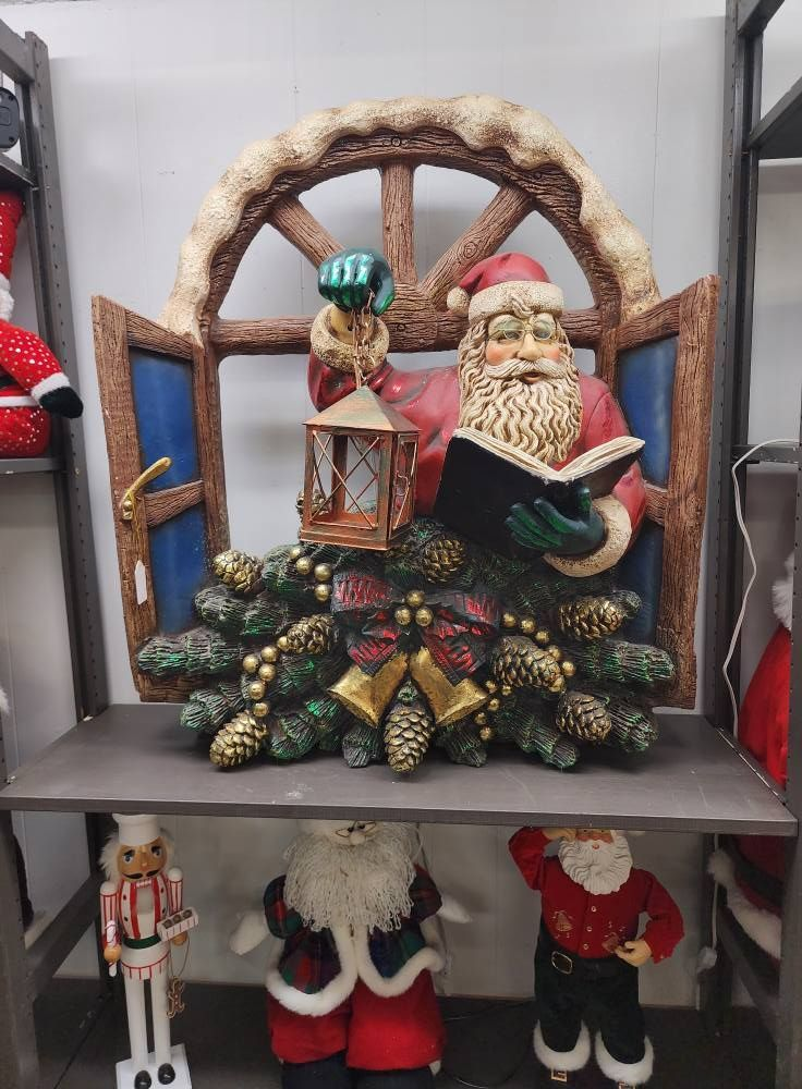 Santa Claus decoration reading a book, framed by a window, with festive greenery and bells.