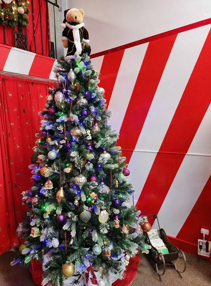 Christmas tree decorated with ornaments, topped with a teddy bear, set against a red and white striped background.