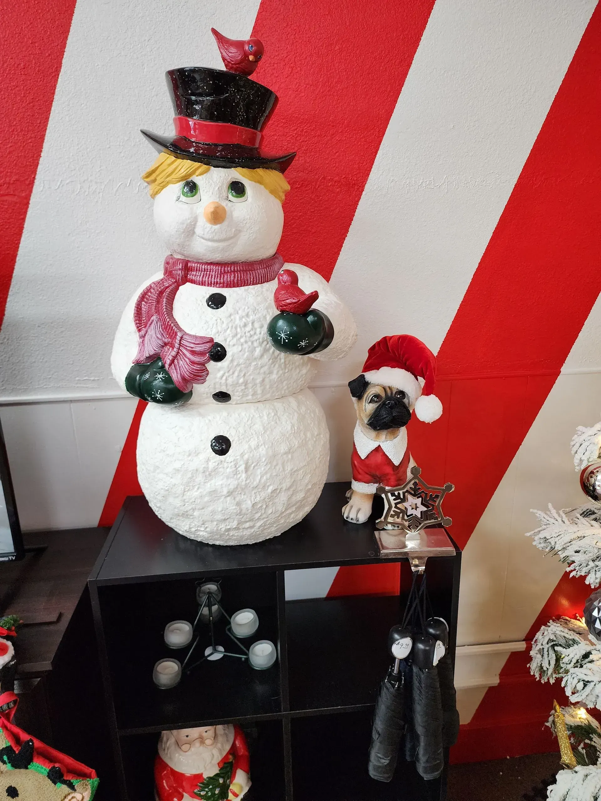 Snowman decoration with red scarf and bird, next to a pug in a Santa hat, on a black shelf.