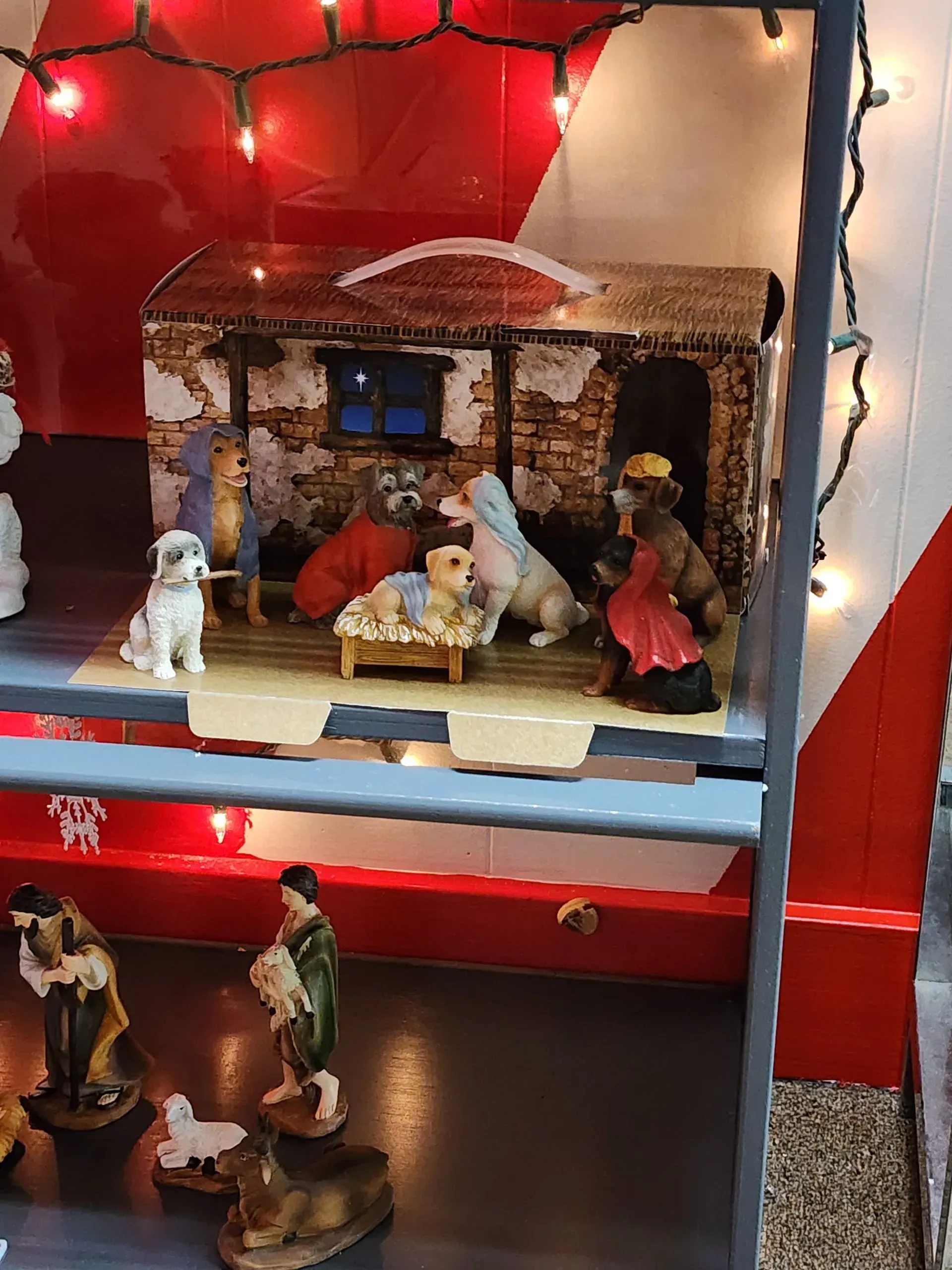 Nativity scene with dog figures; red and white backdrop, string lights.