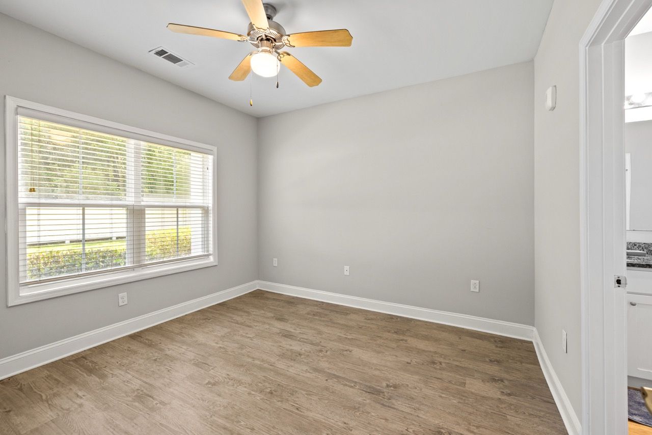 Empty apartment bedroom with large window, blinds, gray walls, wood-look flooring, and a ceiling fan.