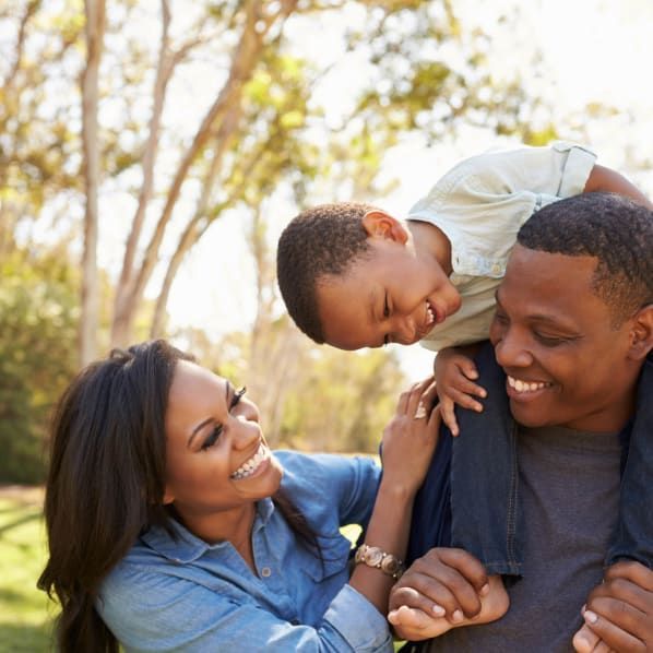 A smiling family enjoys an outdoor setting, with a child sitting on a parent's shoulders while the other parent watches.