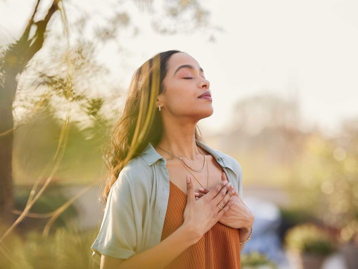 Woman standing outdoors in warm sunlight, eyes closed, hands on chest, with trees softly blurred behind her Woman standing outdoors in warm sunlight, eyes closed, hands on chest, with trees softly blurred behind her