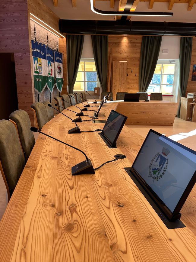 Sala conferenze con tavolo in legno curvo, computer portatili e microfoni di fronte a una parete finestrata.