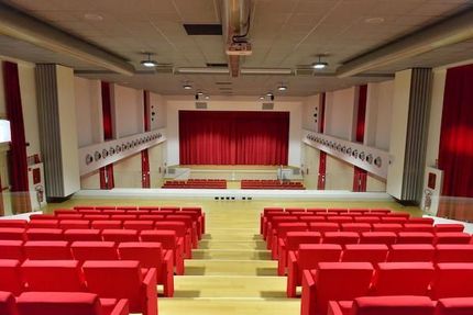 Auditorium vuoto con posti a sedere, palco e tende rossi; illuminazione neutra.