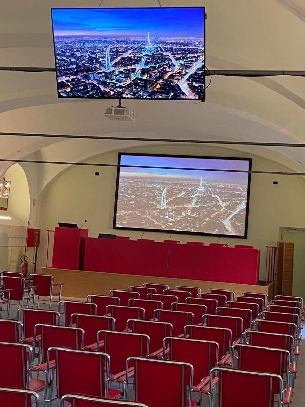 Auditorium con sedie rosse e podio, due schermi che mostrano una vista aerea notturna di una città.