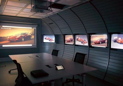 Sala conferenze con un grande tavolo, più schermi che mostrano un'auto rossa e un proiettore.