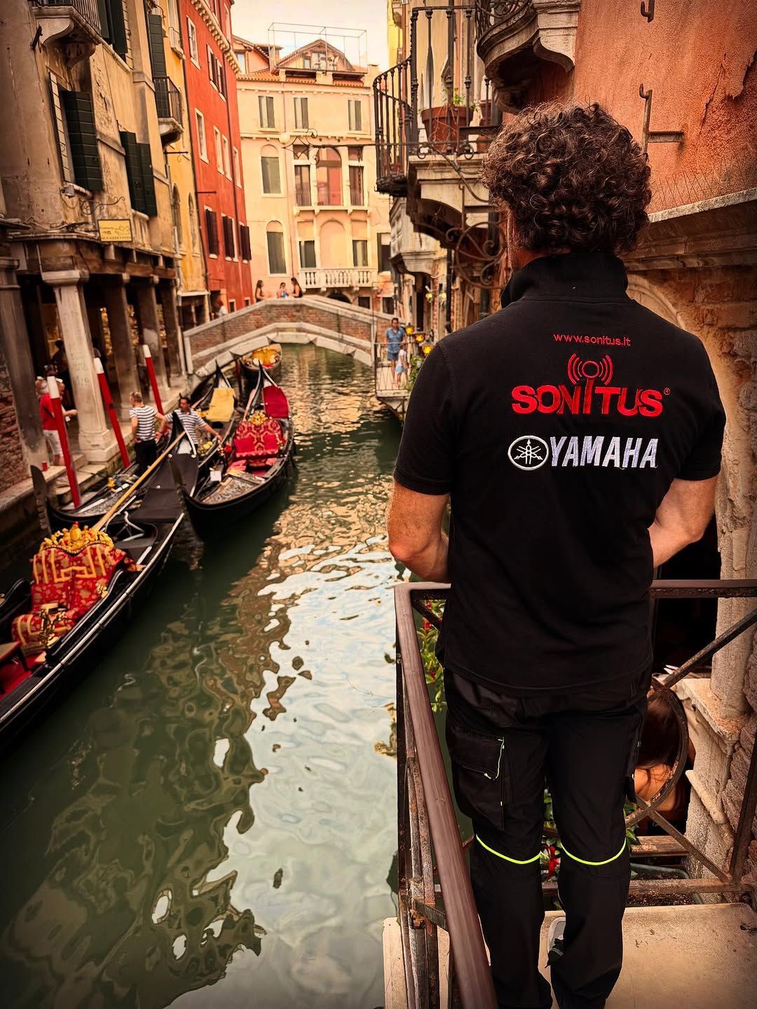 Uomo con camicia nera con logo che si affaccia sul canale veneziano con le gondole.