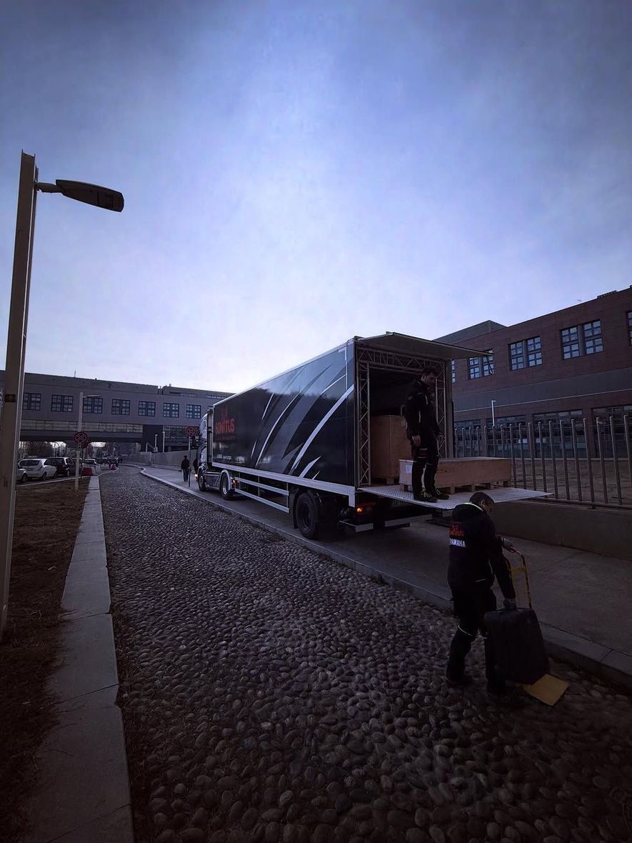 Un camion scarica rifornimenti all'ingresso di un edificio. Una persona trasporta bagagli. Nuvoloso, luce diurna fresca.