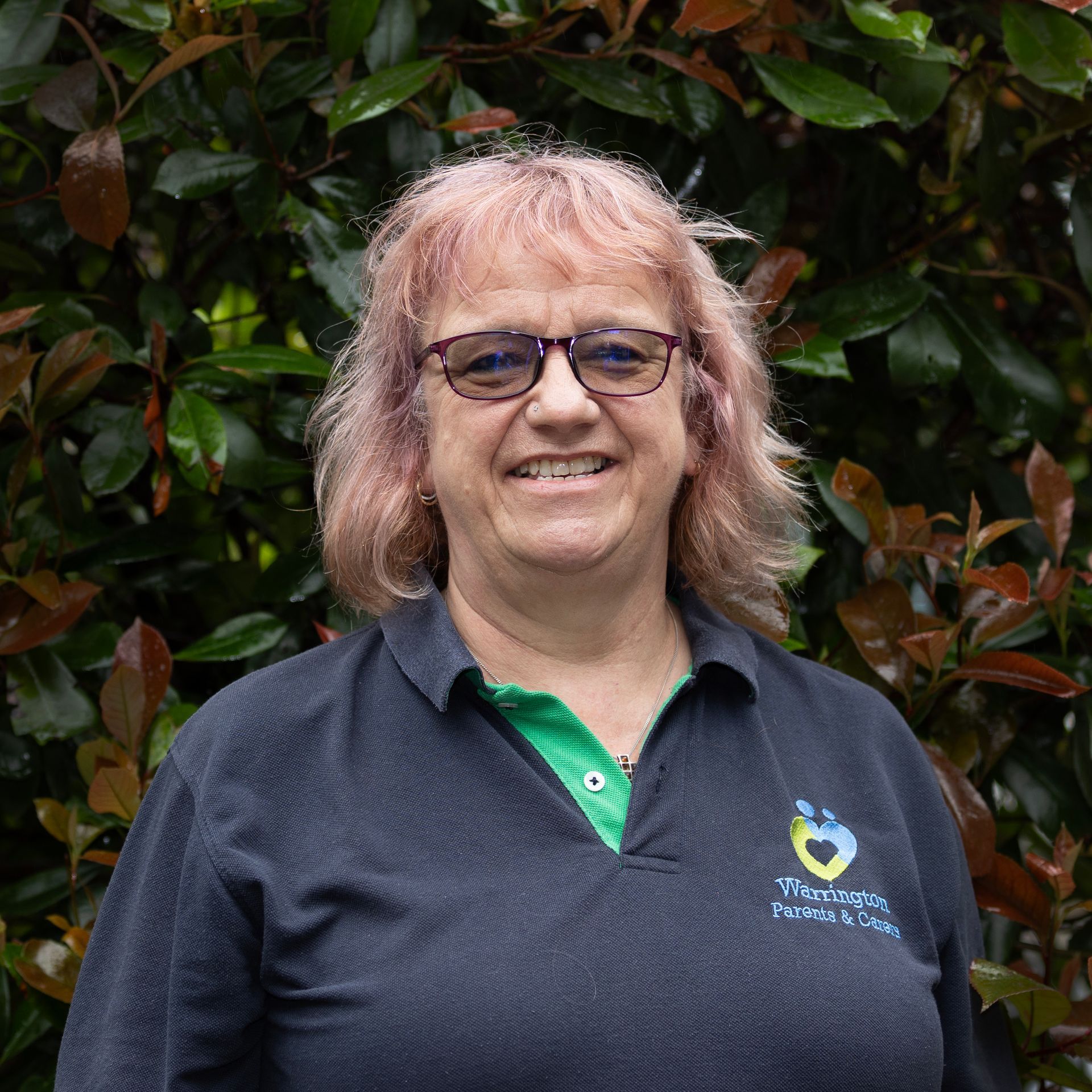 A woman wearing glasses and a black shirt is smiling in front of a bush.