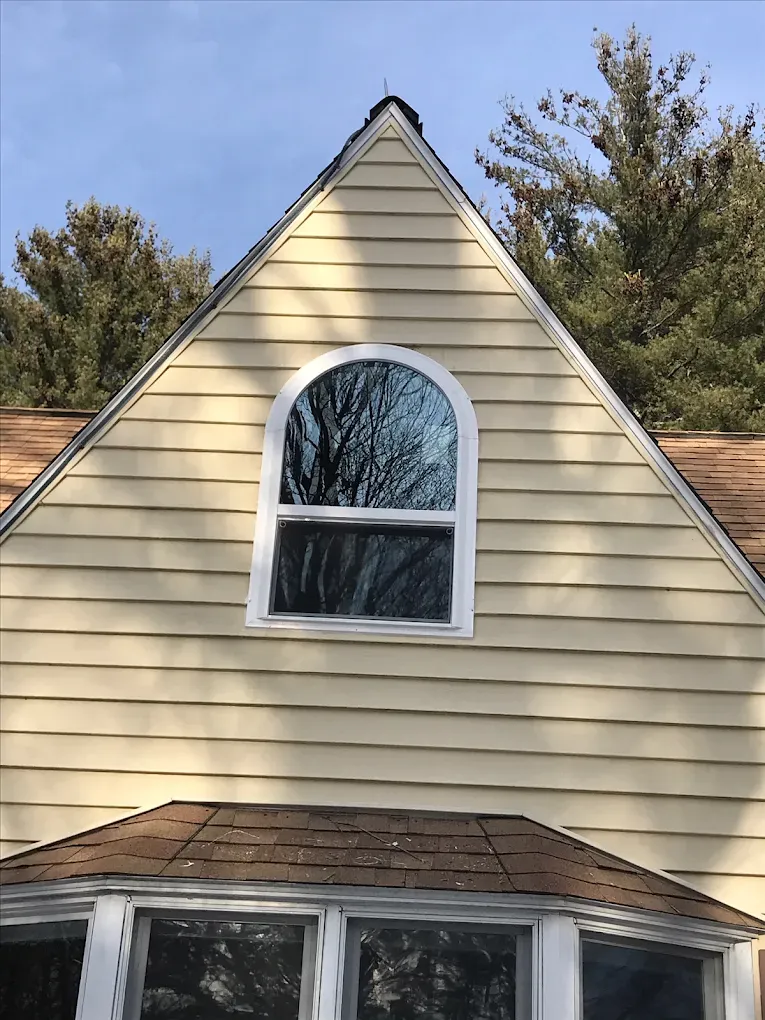Front of a beige house with a steep gable and an arched second-story window beneath a blue sky