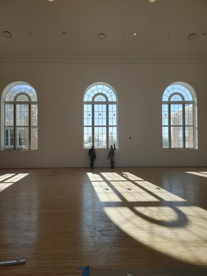 Large sunlit hall with tall arched windows, two people near the center, and long shadows on the wooden floor