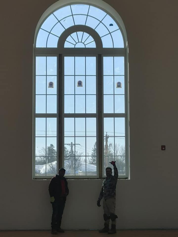 Two people stand by a tall arched window in a bright gallery, looking outside.