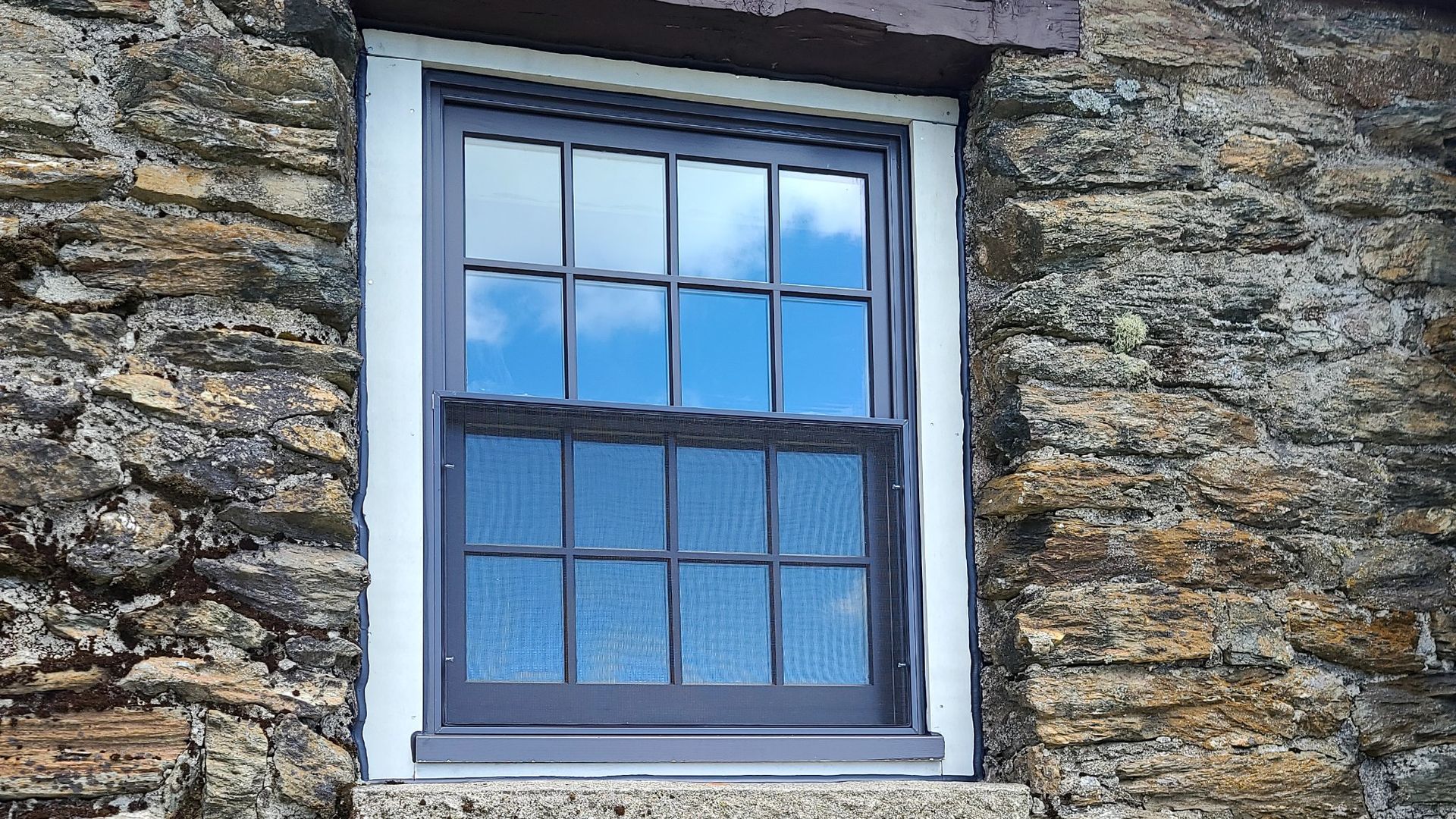 Blue-framed window set in a rough stone wall, with reflected sky and closed curtains behind the glass.