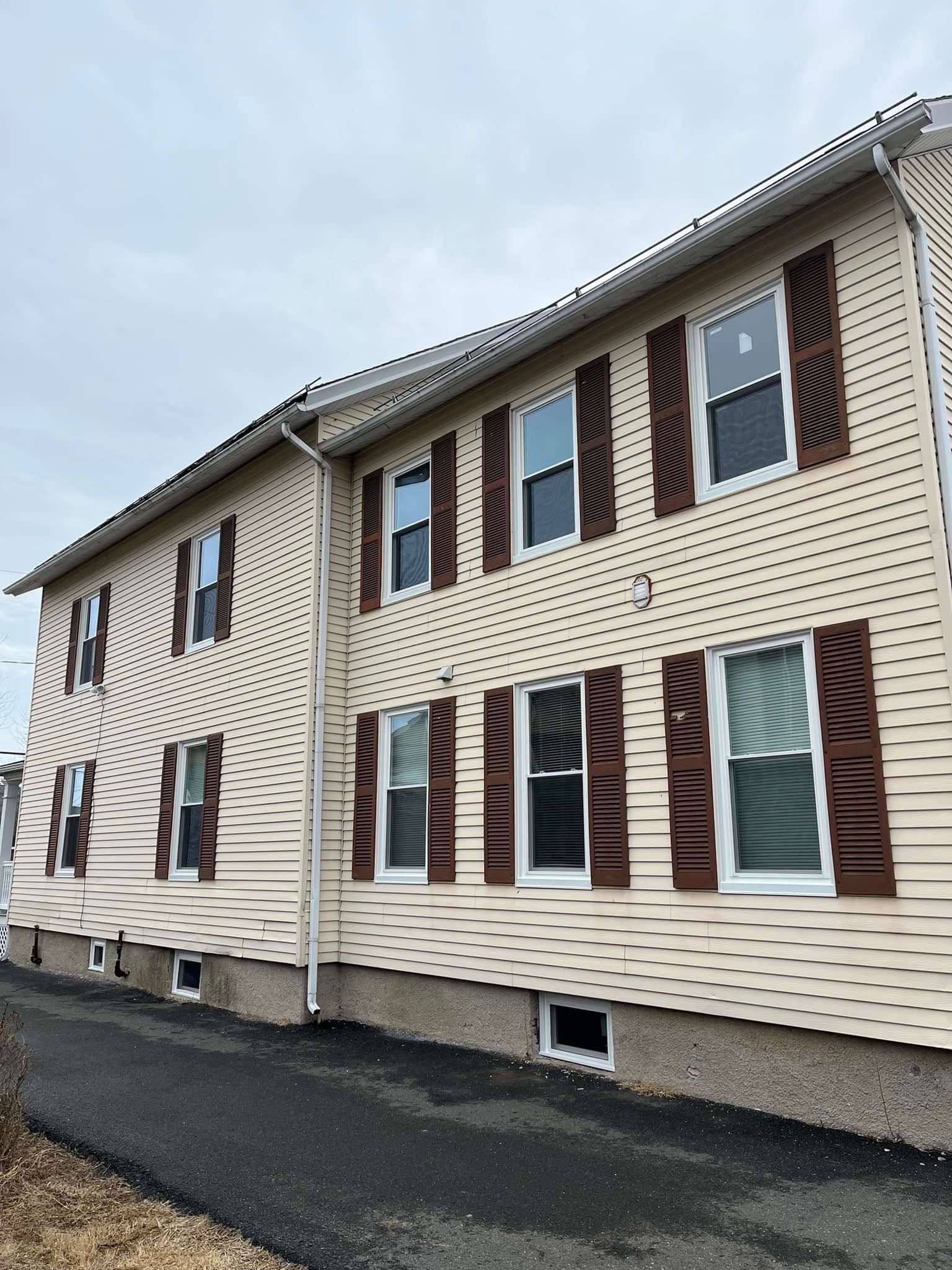 Two-story beige apartment building with brown shutters beside a paved road under a cloudy sky