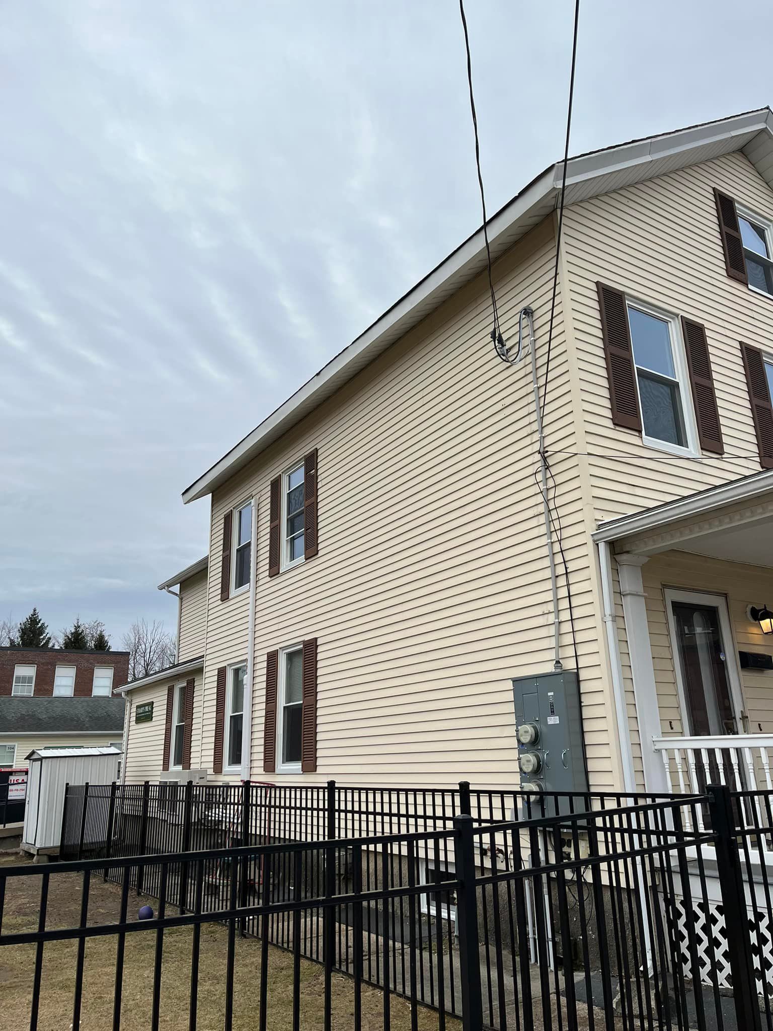Residential house with beige siding, dark shutters, and a black metal fence under a cloudy sky