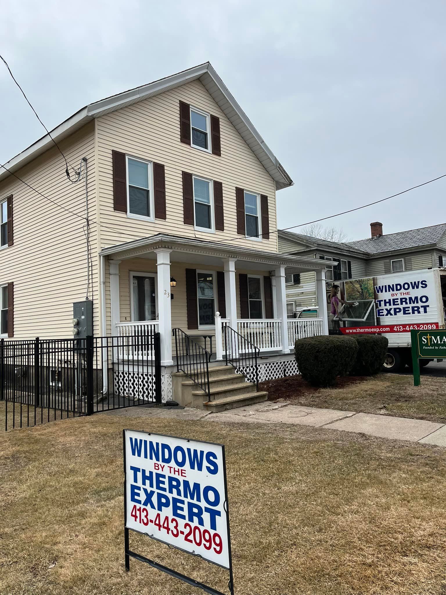 Two-story tan house with dark shutters and a Windows Thermo Experts sign in the front yard.