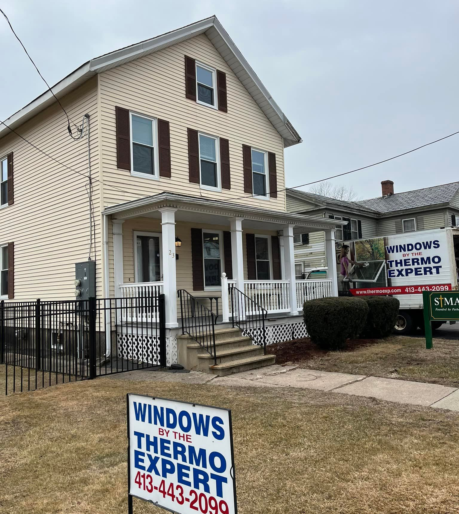 Two-story beige house with black shutters and white porch, with Windows Thermo Expert signs in front yard