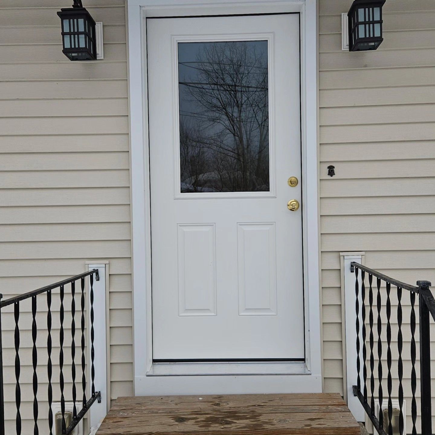 Front porch with a white door, beige siding, black railings, and wall lanterns.