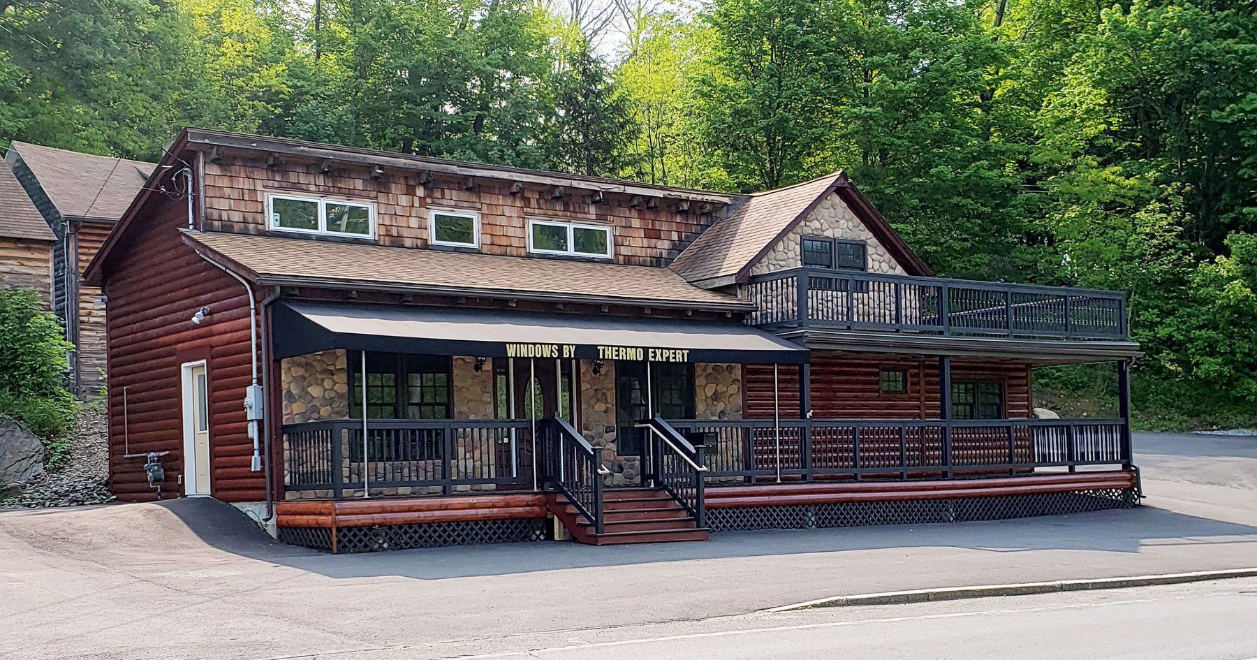 Rustic red lodge with porch and black railings beside a gravel lot, surrounded by trees