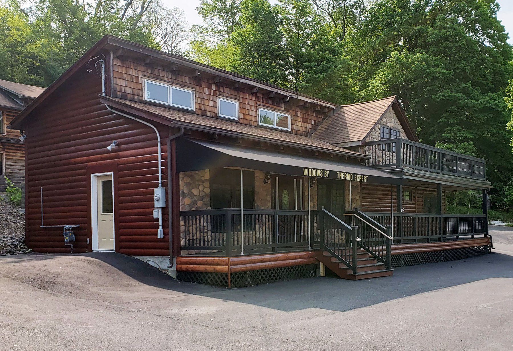 Rustic log cabin with wraparound porch and deck, surrounded by trees and a gravel driveway