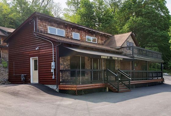 Rustic log cabin with wraparound porch and deck, surrounded by trees and a gravel driveway