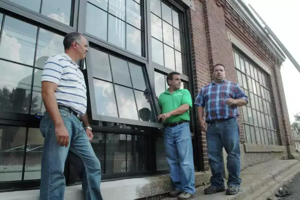 Three men standing outside a brick building with large windows, talking on the sidewalk.