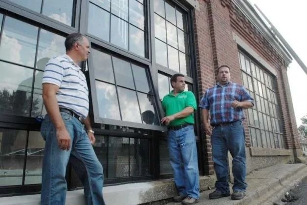 Three men stand outside a brick building by large windows, talking on a sidewalk.