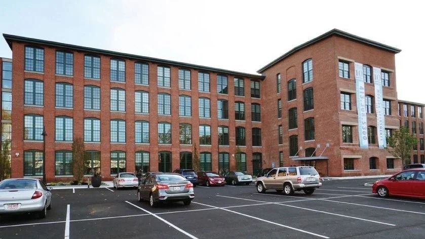 Brick apartment building with parked cars in a lot under an overcast sky