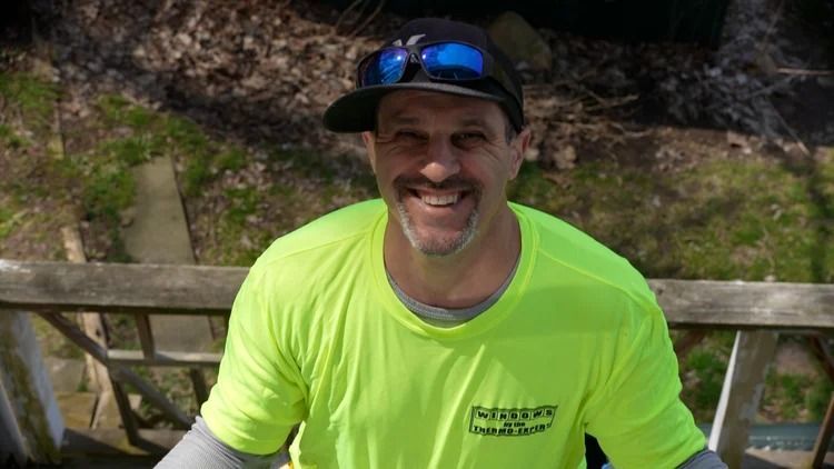 Smiling man in neon yellow shirt and black cap sitting outdoors by a wooden fence
