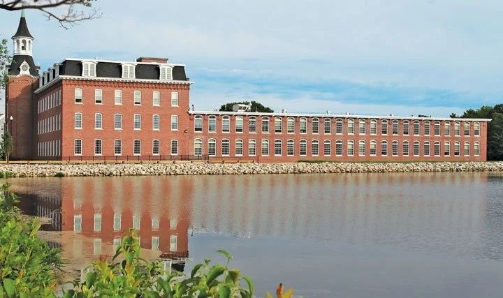 Long brick industrial building beside calm water, with reflections and a cloudy sky