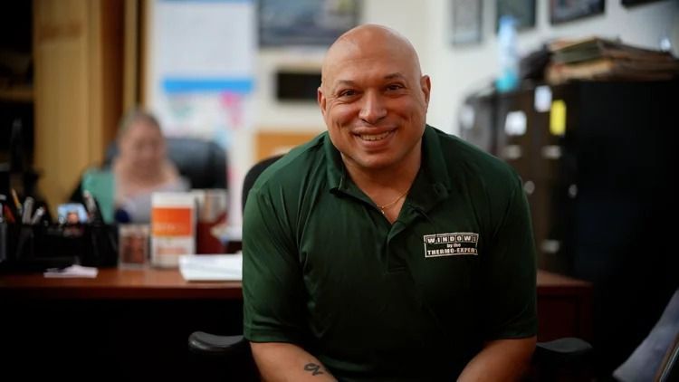 Smiling man in a green polo shirt seated indoors at a counter with blurred people and posters behind him