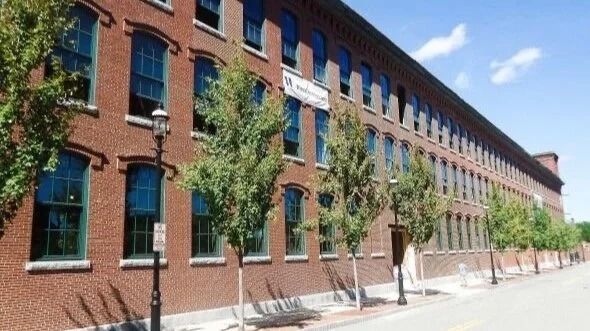 Red brick industrial-style building with rows of windows and young trees along a sunny sidewalk.