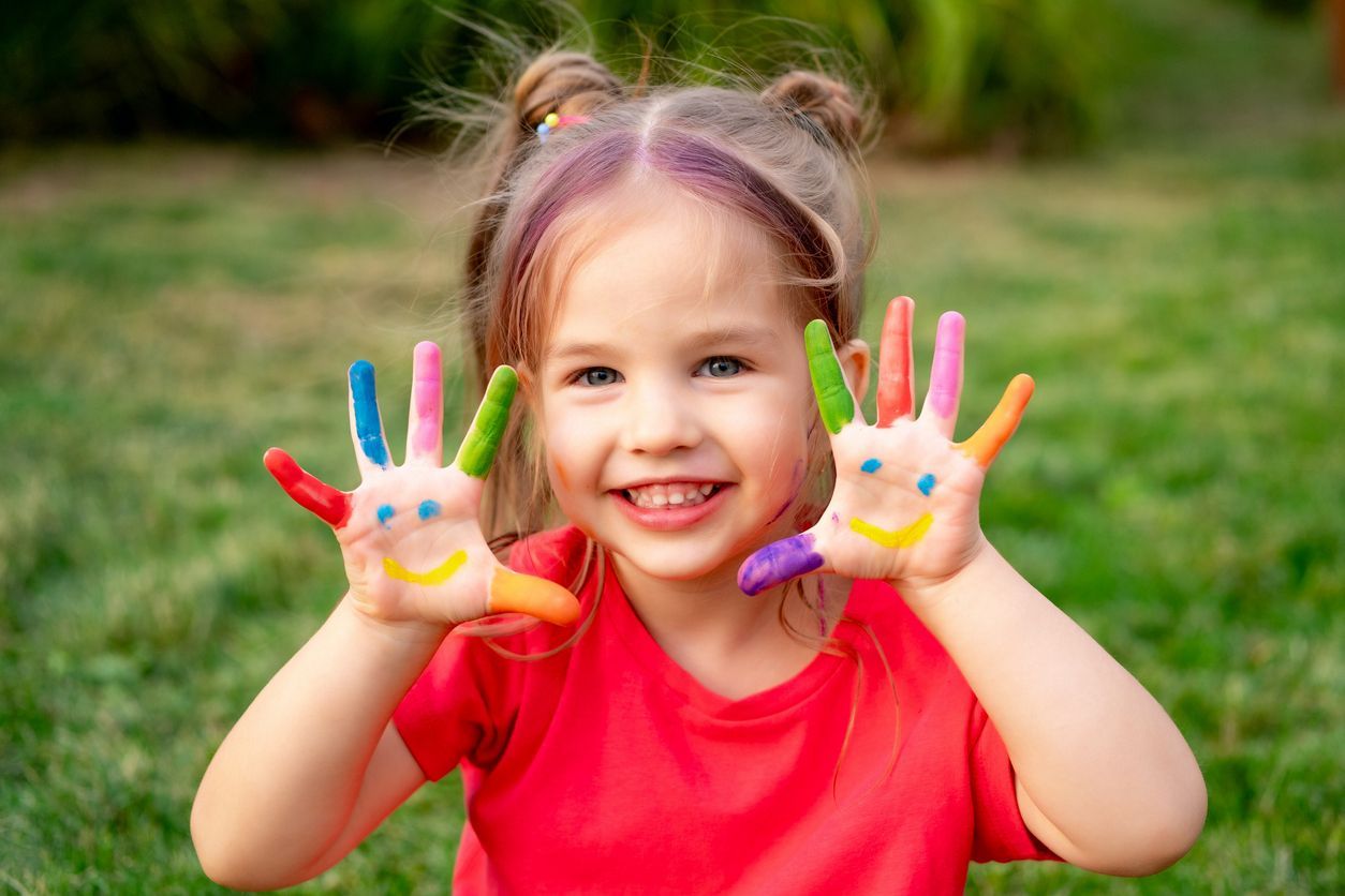 Girl with painted hands displaying smiley faces, smiling in a grassy outdoor setting.