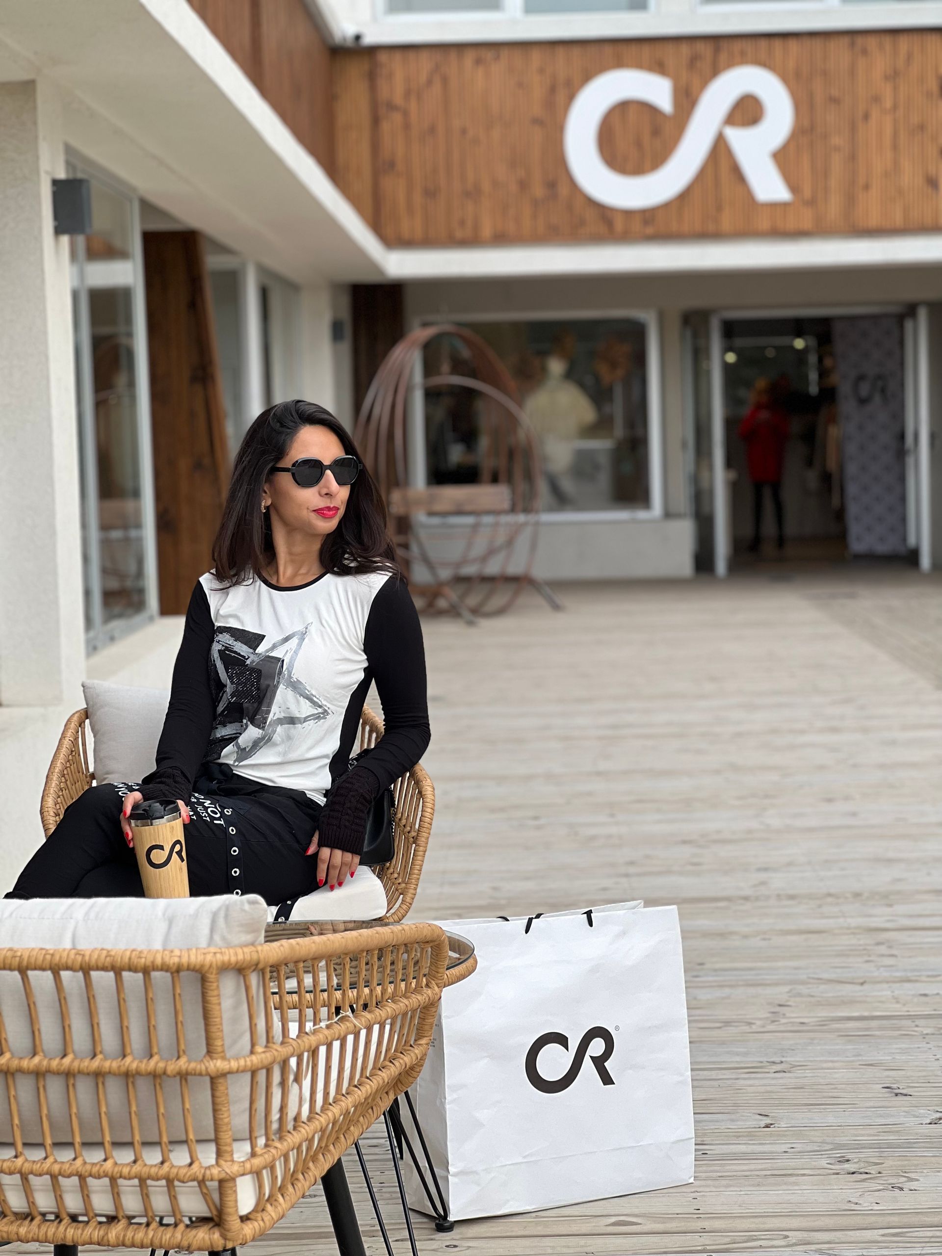 Mujer con gafas de sol sentada afuera de una tienda sosteniendo una taza de café y una bolsa de compras con el logotipo 