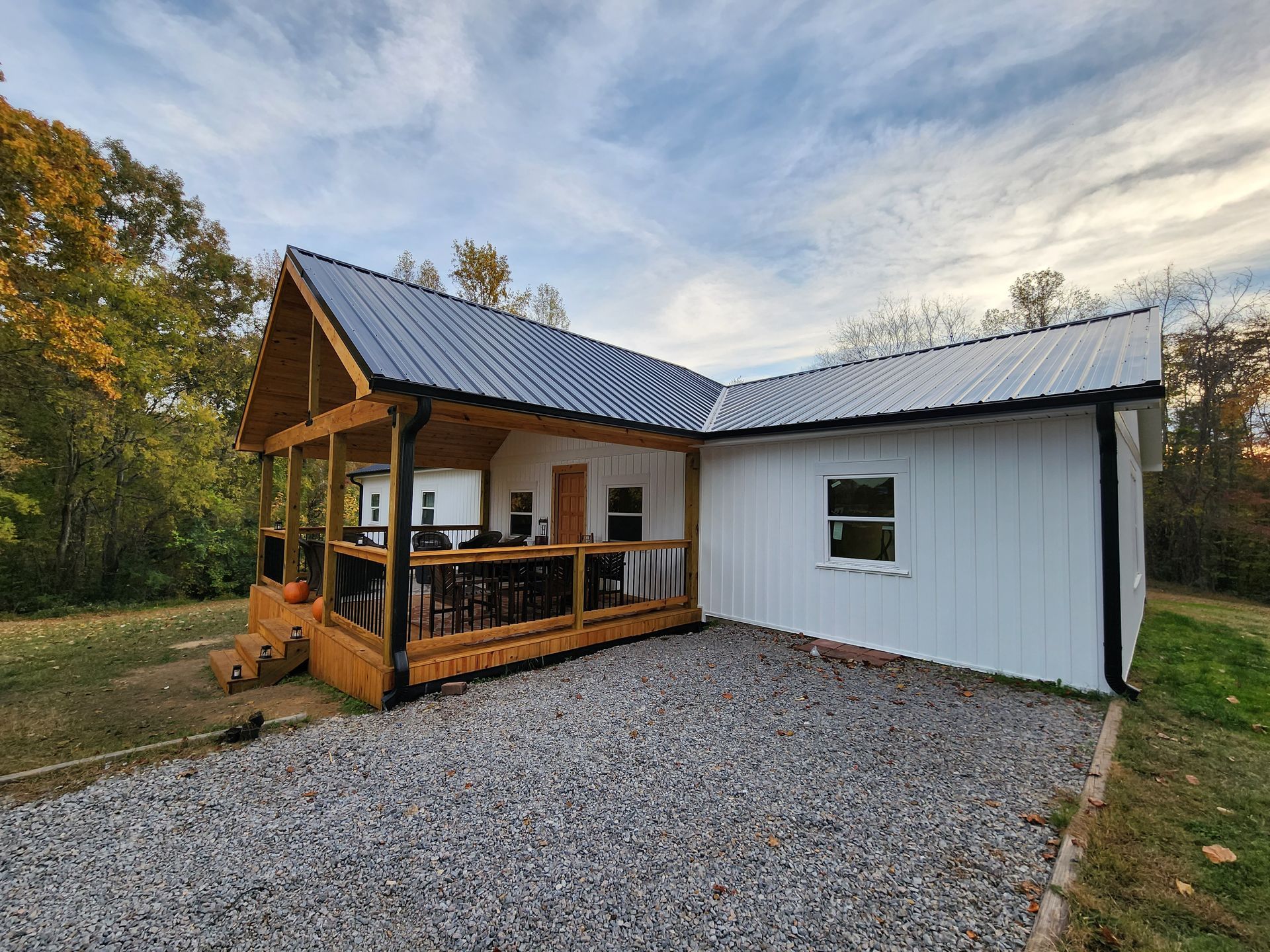 White cabin with dark roof and porch, gravel driveway, surrounded by trees.