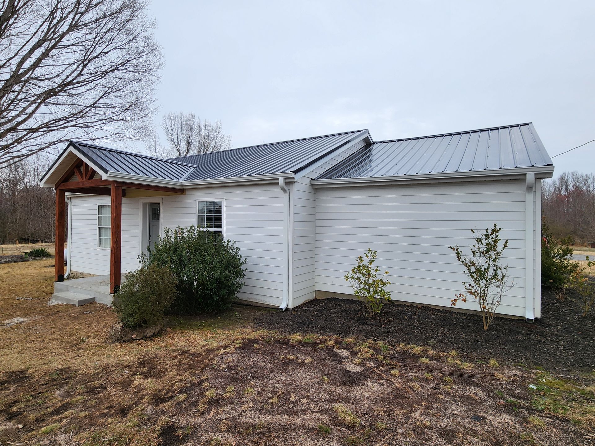 White house with metal roof, small porch, and some bushes on a cloudy day.