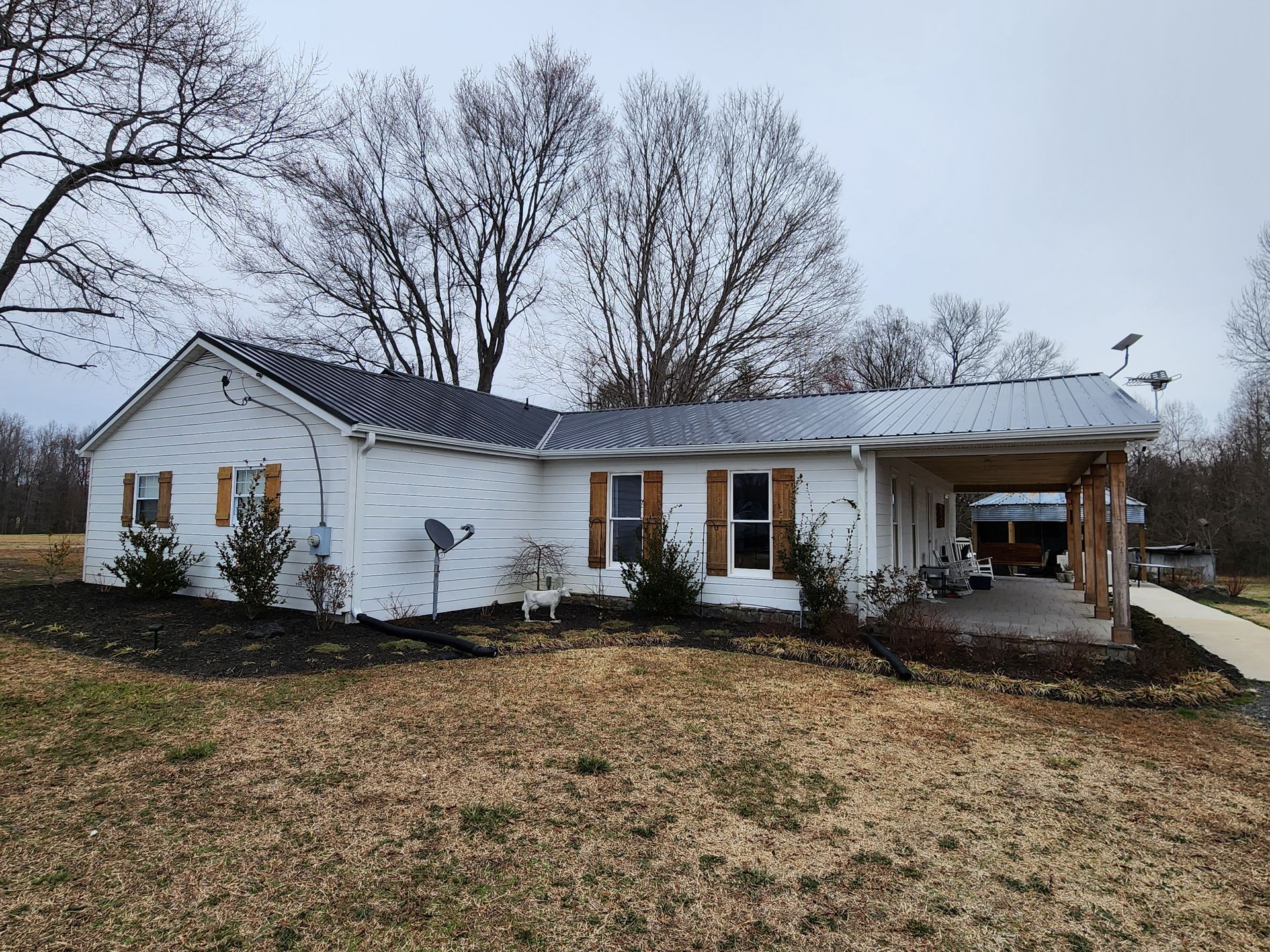 White house with metal roof and carport on a cloudy day. Brown shutters, leafless trees, and brown grass in the yard.